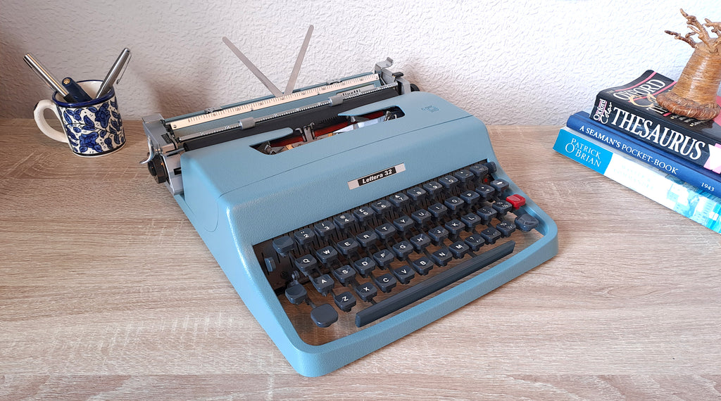 A blue vintage Olivetti Lettera 32 typewriter sits on a light wood desk.