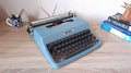 A blue vintage Olivetti Lettera 32 typewriter sits on a light wood desk.